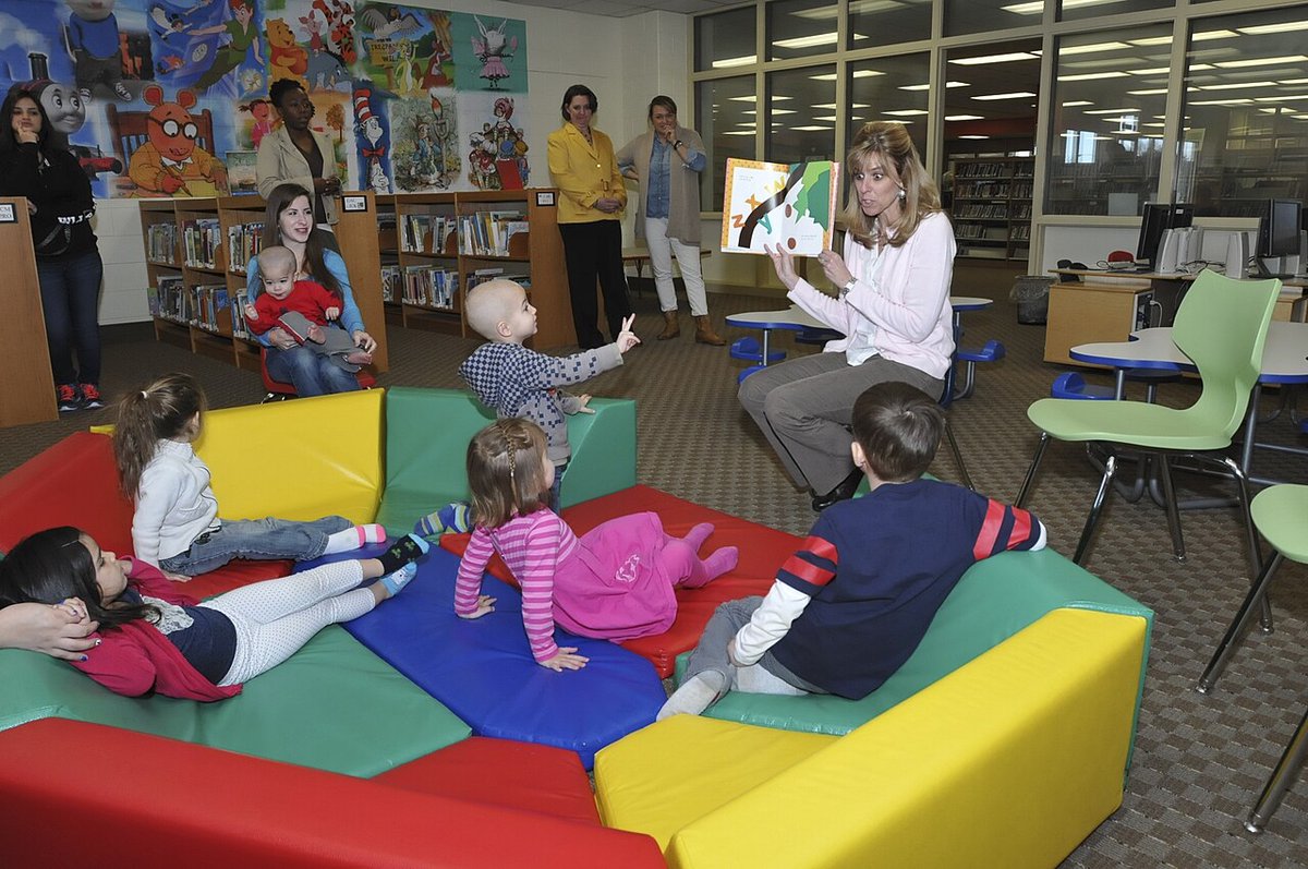 A reader holds up a picture book for a handful of young kids sprawled on bright foam cushions in a children's library corner
