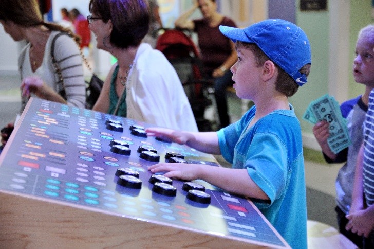A boy in a blue cap presses the big round buttons on a light-up control board in the amphitheater at Pretend City Children's Museum