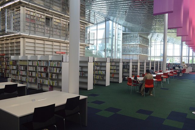 A bright public library interior with long rows of low bookshelves and reading tables, with no one in view