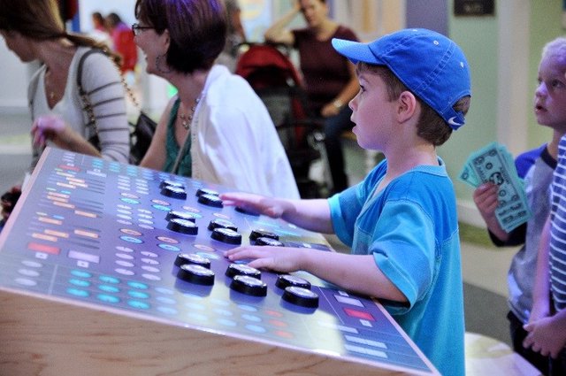 A boy in a blue cap presses the big round buttons on a light-up control board in the amphitheater at Pretend City Children's Museum