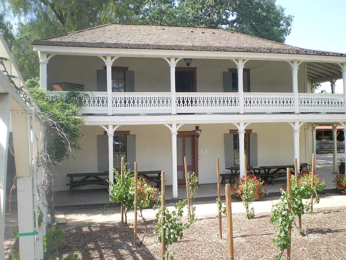 Two-story white adobe house with a wraparound upper balcony, picnic tables and young grapevines in the foreground