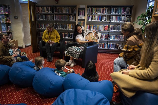 A reader in a library holds up an open picture book for toddlers sitting on blue beanbags while parents and a librarian watch