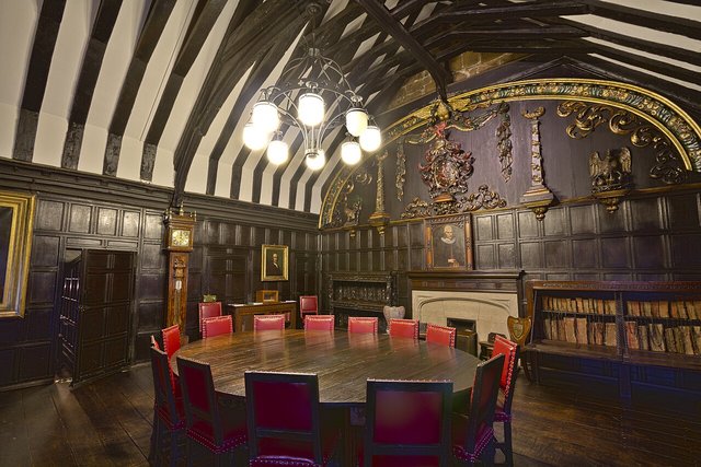 A historic wood-panelled library reading room with a round table and red chairs under a timbered ceiling