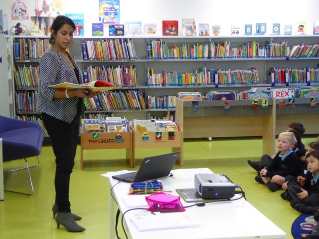 A reader holding up an open picture book in a children's library while young kids sit on the floor listening, with colorful shelves of kids' books behind her