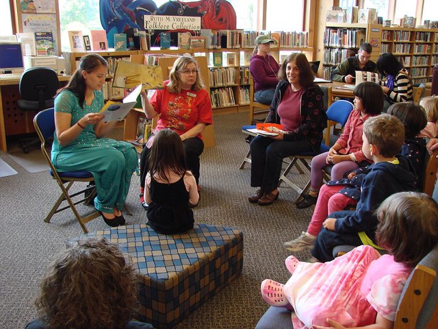 Two librarians reading picture books aloud to a circle of young children seated on chairs and a cushion in a library children's section