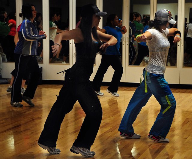 Dancers in sneakers and loose track pants doing a Zumba routine on a wood studio floor in front of a mirrored wall