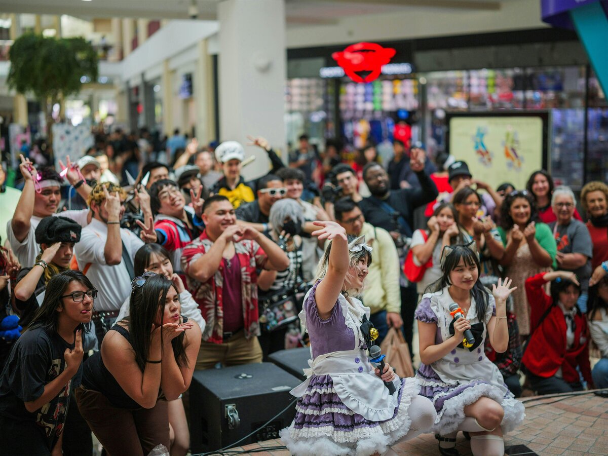 Crowd in a shopping mall cheering around two performers in lavender maid-style outfits holding microphones