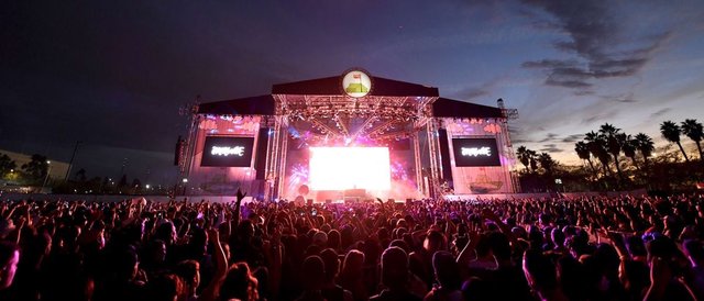 Crowd facing a lit festival stage at dusk with palm trees on the right