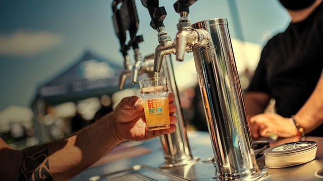 A festival-goer holds a Summit Beer Festival tasting glass under a chrome tap tower as a golden pour fills it on a sunny afternoon
