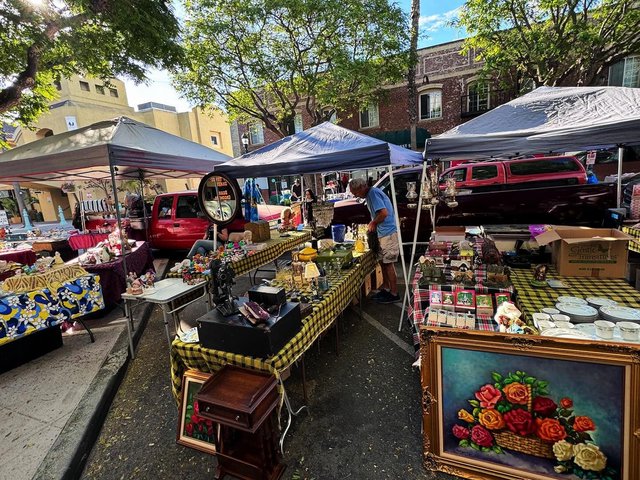 Antique vendor tables under canopies on a downtown Torrance street, with a framed rose painting, vintage projector and china on display