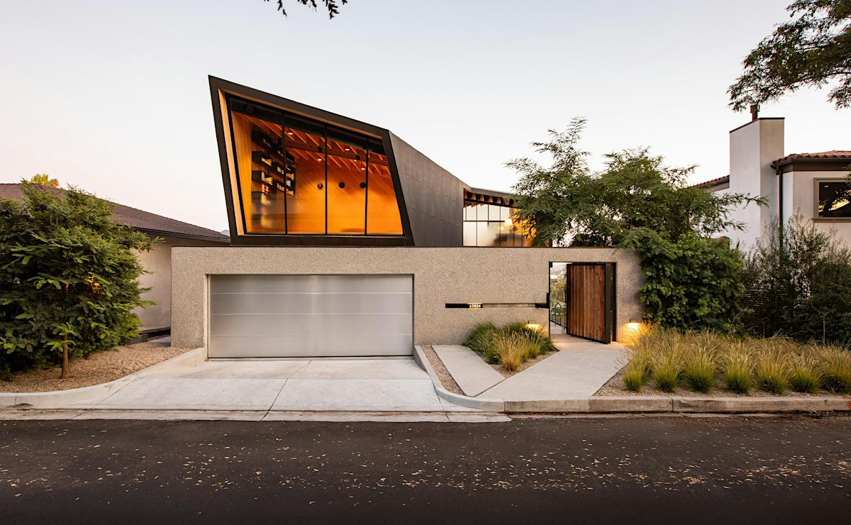 Modern home with an angular glass window glowing amber above a concrete garage wall and wooden gate at dusk