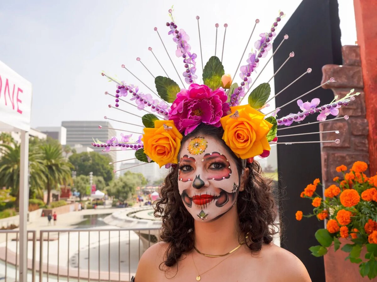 Woman in sugar skull face paint and a headdress of orange and magenta flowers, with marigolds and the Grand Park fountain behind her