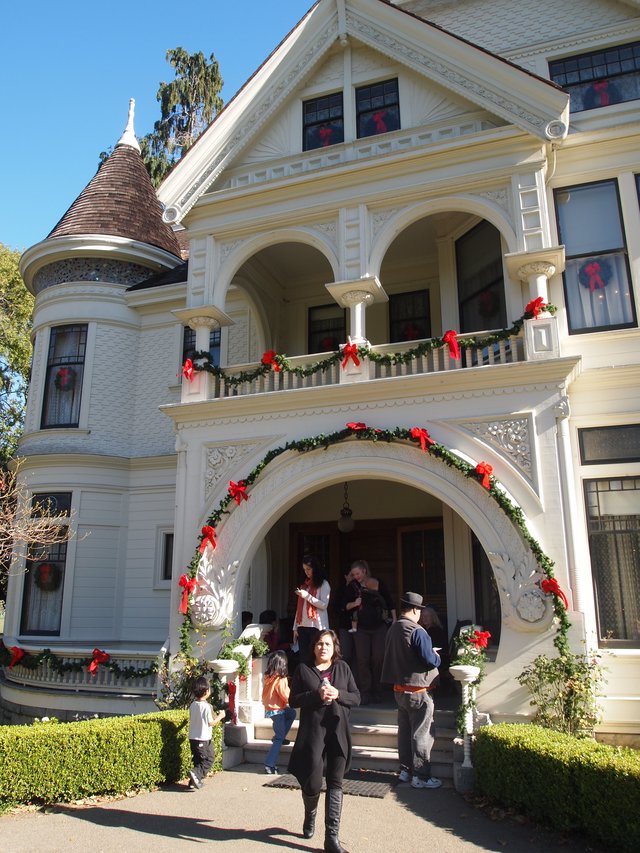 A white Victorian farmhouse porch decorated for a fall community gathering, with visitors walking past out front