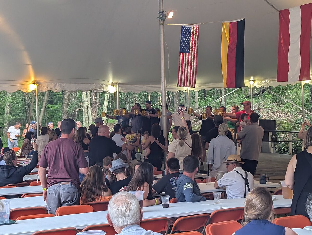 Contestants on a tented stage hold full beer steins at arm's length while a seated crowd watches a stein holding competition