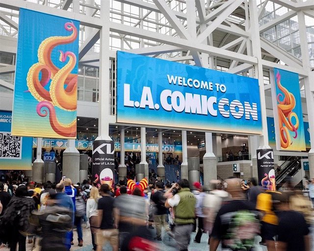 Crowds walking beneath a giant Welcome to L.A. Comic Con screen in the glass lobby of the Los Angeles Convention Center, with orange tentacle banners on either side