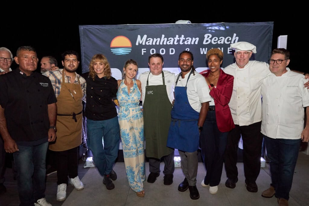 Chefs in aprons and whites pose with guests in front of the Manhattan Beach Food & Wine banner at night
