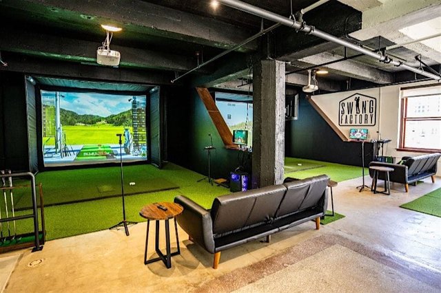 Indoor golf simulator bays with projected fairways, green turf and lounge couches inside Swing Factory Golf