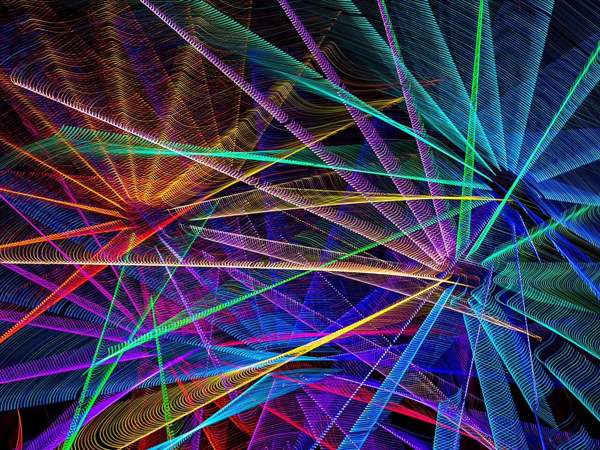 Long-exposure photo of a ferris wheel with rainbow light trails radiating from its spokes against a night sky