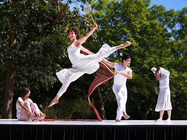 Dancers in white costumes perform on an outdoor stage under tall trees, one leaping mid-air holding a badminton racket as a red net trails behind
