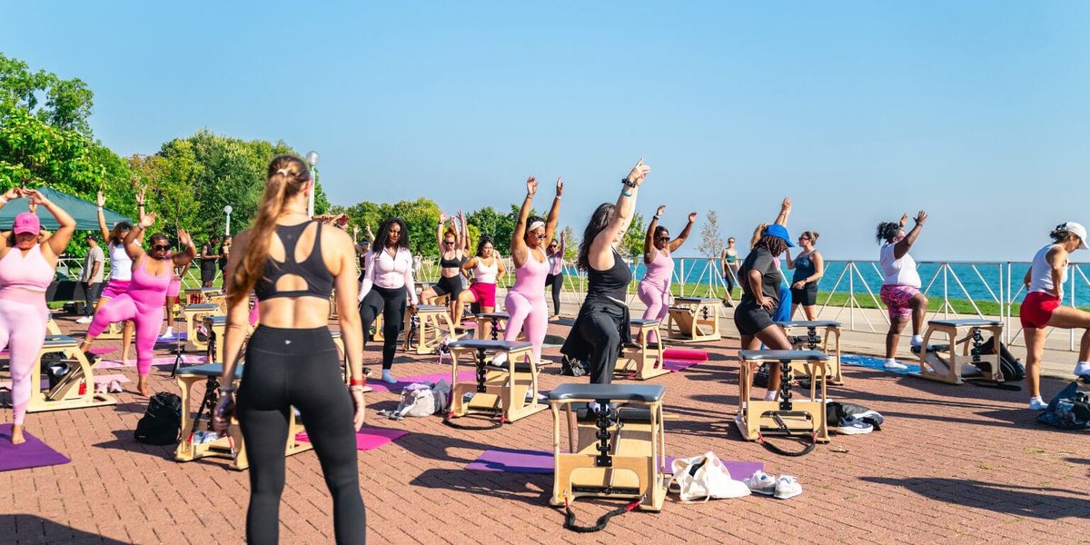 Group doing a raised-arm stretch on Pilates chairs and mats on a brick plaza beside a lake