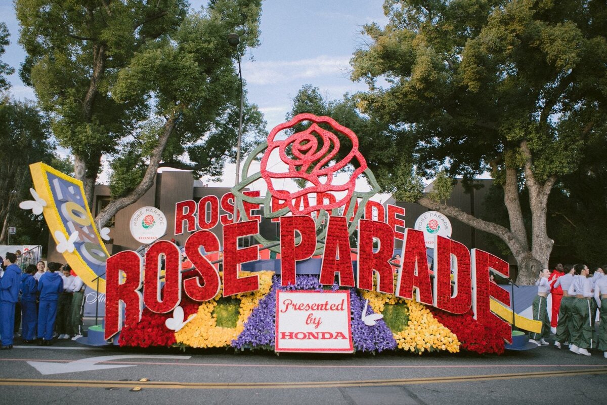 A red, yellow and purple flower-covered Rose Parade float with large lettering and a giant rose, parked on a tree-lined Pasadena street