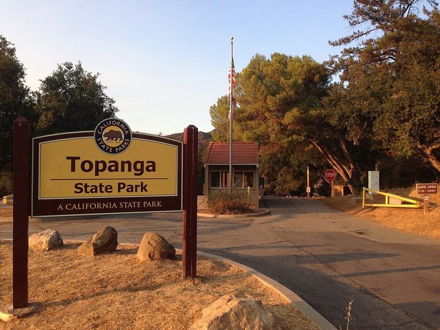 Topanga State Park entrance sign beside a ranger kiosk and flagpole, framed by oak and pine trees