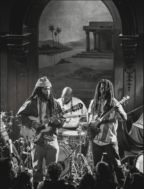 Black-and-white photo of a guitarist, drummer and bassist performing on a small stage under an arched mural