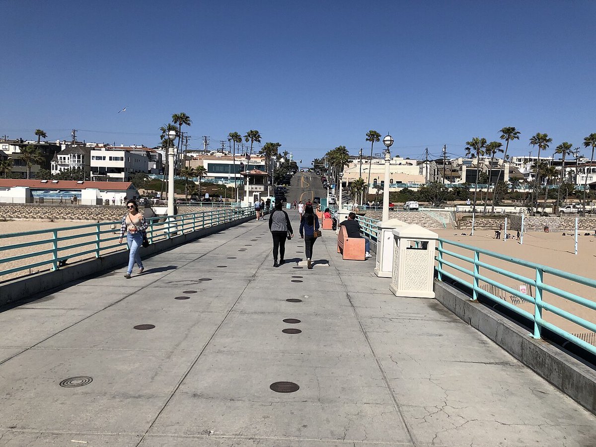 People walking along Manhattan Beach Pier toward the palm-lined street that climbs into downtown Manhattan Beach