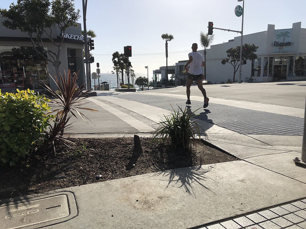 A runner crosses a downtown Manhattan Beach street with palm trees and the Pacific Ocean visible at the end of the block