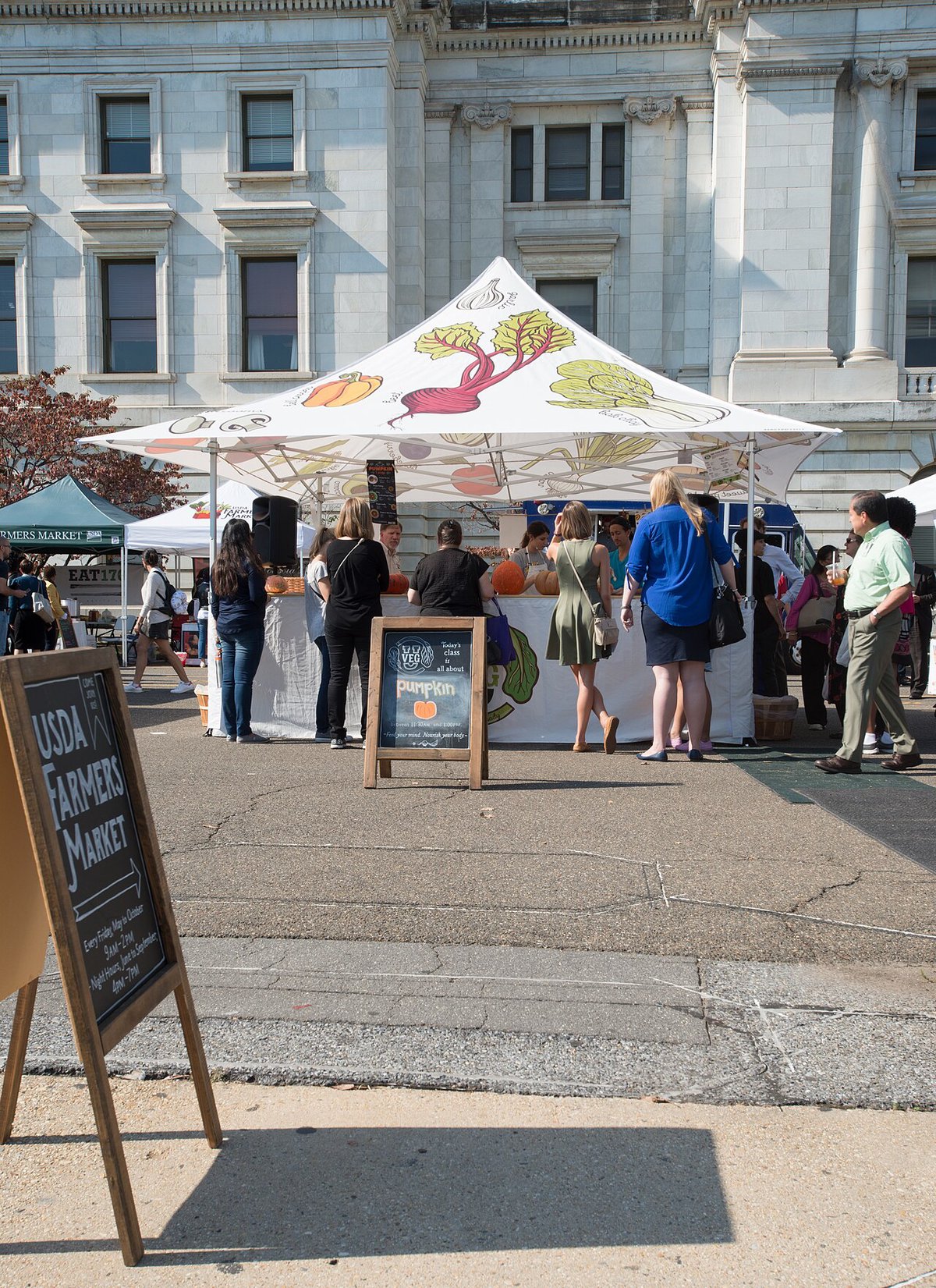 Shoppers lined up at a white canopy stall with illustrated vegetables and a pumpkin sign, beside a chalkboard farmers market sign