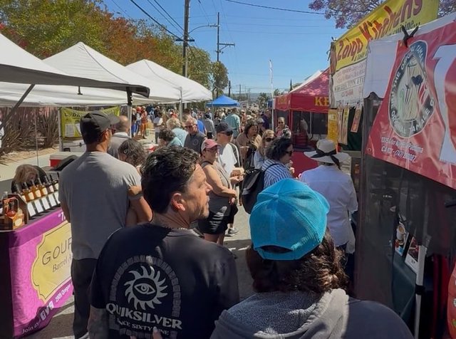 Shoppers crowding a closed street between white vendor tents and food stalls at the North Redondo Beach Farmers Market