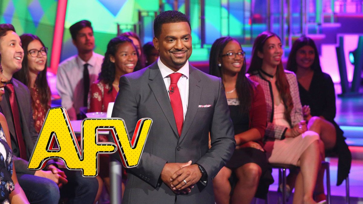 Host Alfonso Ribeiro smiling in a gray suit and red tie on the America's Funniest Home Videos set, with seated audience members behind him and the yellow AFV logo in the corner
