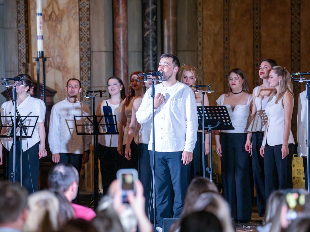 A soloist sings into a microphone in front of a large choir dressed in white and black, with an audience watching