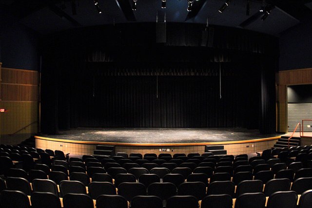 An empty theatre stage with black curtains in front of rows of dark blue seats