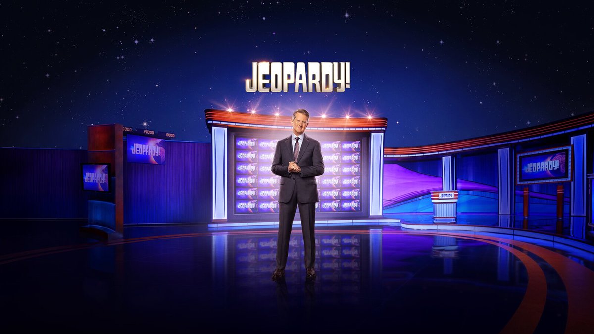 Host Ken Jennings in a gray suit stands on the Jeopardy! stage in front of the game board, under the gold Jeopardy! logo and a starry blue backdrop