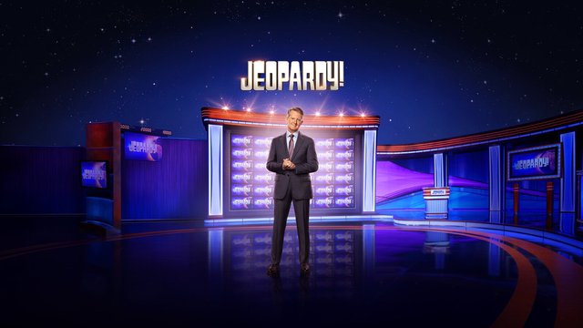Host Ken Jennings in a gray suit stands on the Jeopardy! stage in front of the game board, under the gold Jeopardy! logo and a starry blue backdrop