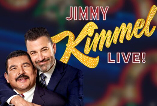 Jimmy Kimmel in a dark suit hugging Guillermo from behind, beside the gold marquee Jimmy Kimmel Live! logo