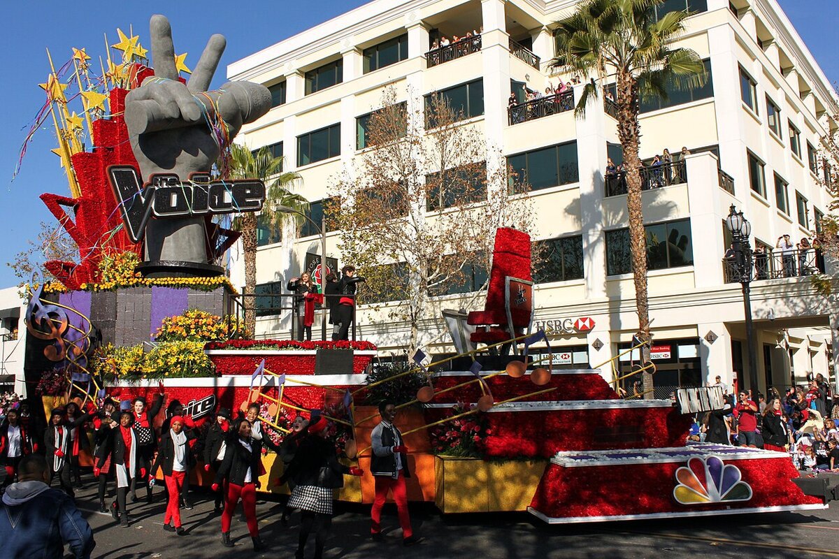 A flower-covered parade float topped by a giant sculpted hand and The Voice logo, with an NBC peacock at the base, rolling down a Pasadena street