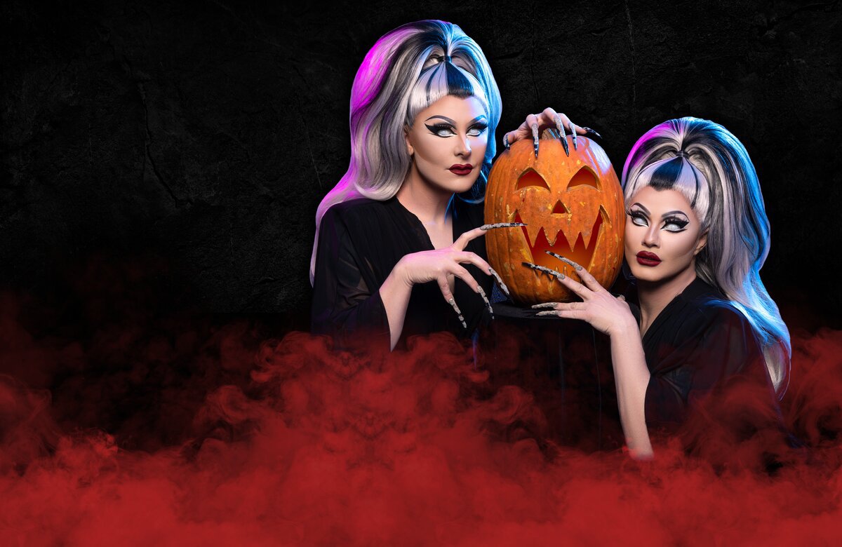The Boulet Brothers in black gowns and long silver nails holding a carved jack-o'-lantern between them, in front of red fog