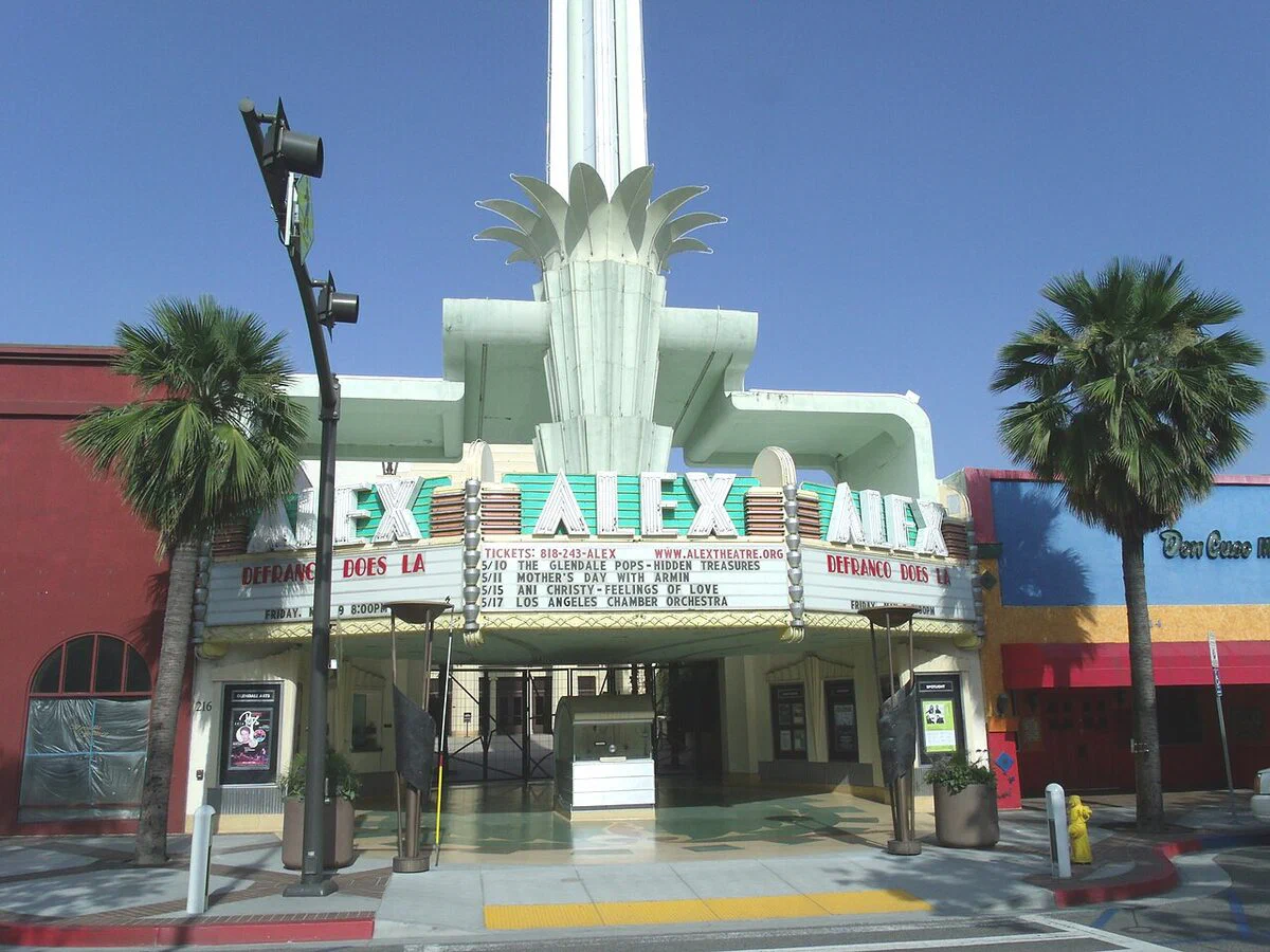 The Alex Theatre's neon ALEX marquee and finned white tower on Brand Boulevard in Glendale on a sunny day, framed by palm trees