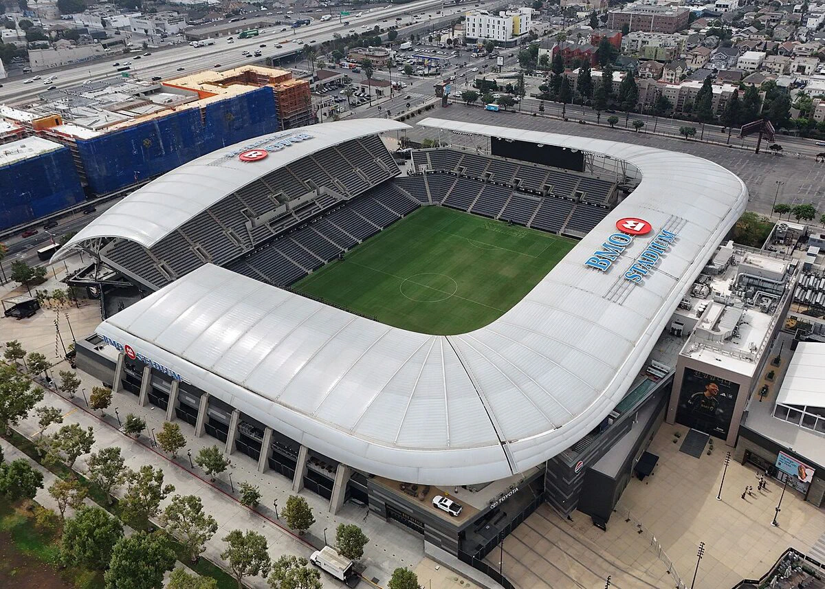 Aerial view of BMO Stadium in Los Angeles, its white roof wrapping a green soccer pitch with the 110 freeway behind