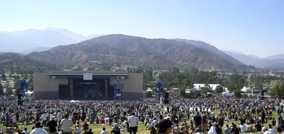 A crowd spread across the lawn of Glen Helen Amphitheater in daylight, facing the covered stage, with dry mountains rising behind it