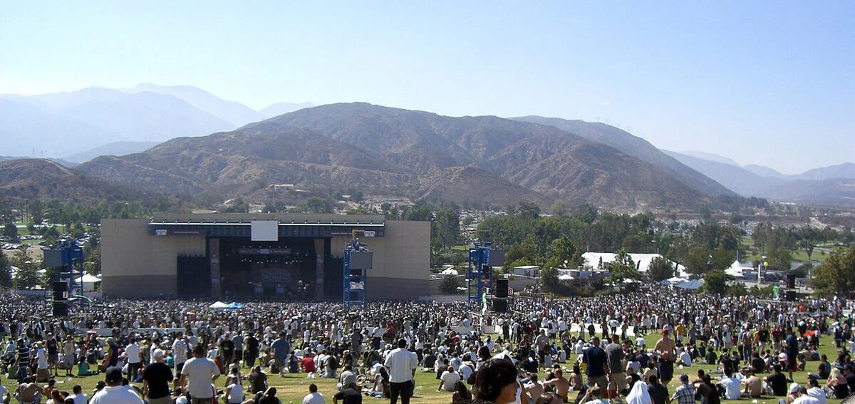 A crowd spread across the lawn of Glen Helen Amphitheater in daylight, facing the covered stage, with dry mountains rising behind it