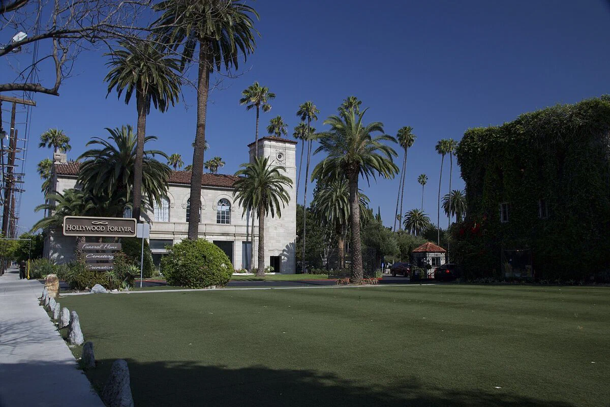 The entrance lawn at Hollywood Forever Cemetery with tall palm trees and a white building beyond the grass