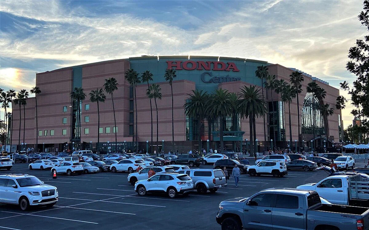 Honda Center's pink stone facade, glass entrance and red Honda Center sign behind palm trees and a full parking lot at sunset