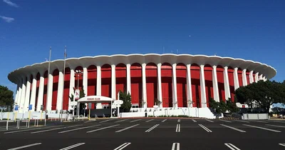 The Kia Forum's ring of tall red columns under a white roof edge, seen across its parking lot in Inglewood on a sunny day