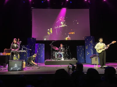 A band playing on the wide stage of The Novo in downtown Los Angeles, with a large video screen and purple lights behind them