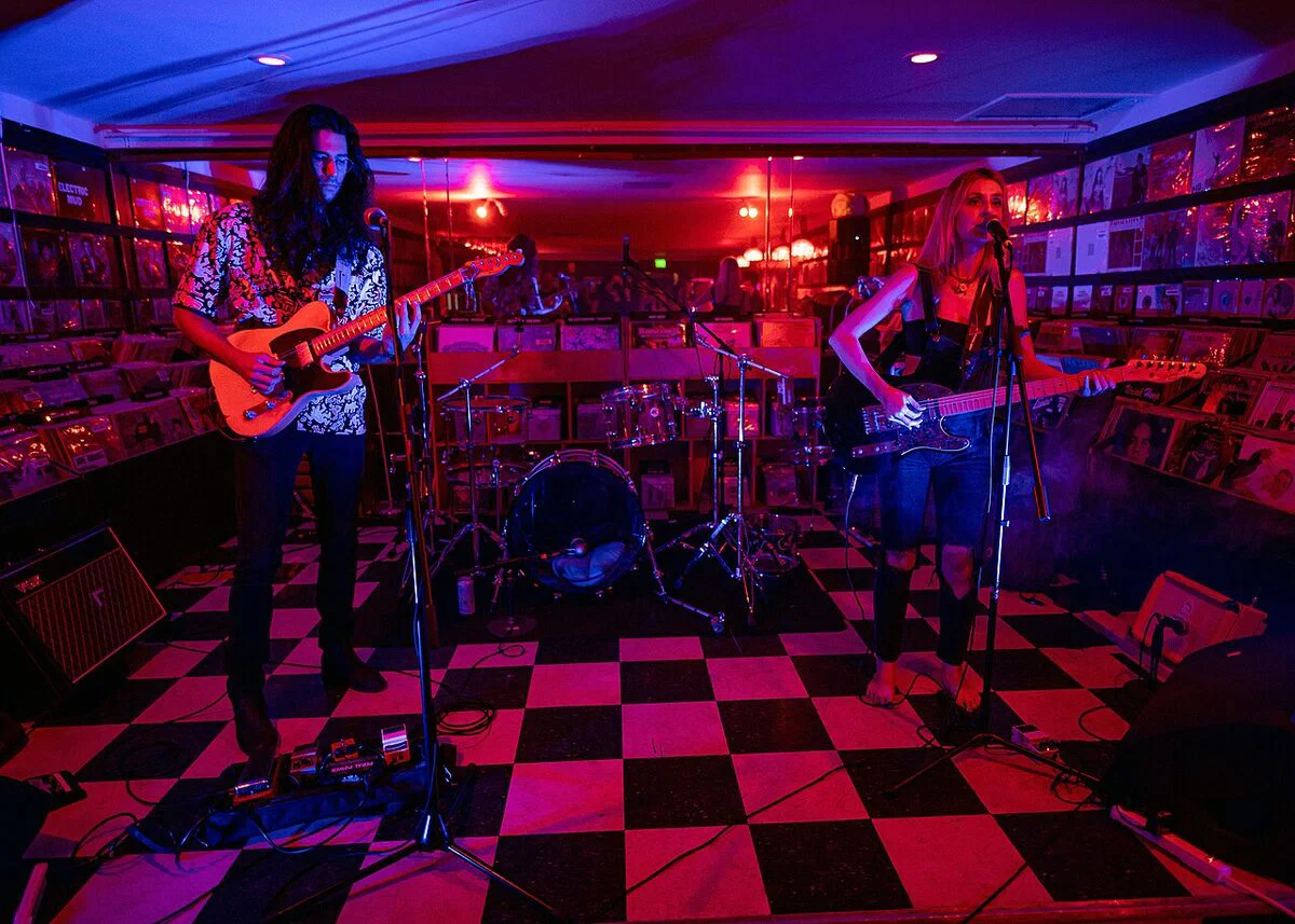 A band playing on the checkered floor at Permanent Records Roadhouse, with walls of record bins on both sides under red and blue light