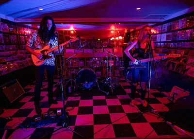 A band playing on the checkered floor at Permanent Records Roadhouse, with walls of record bins on both sides under red and blue light