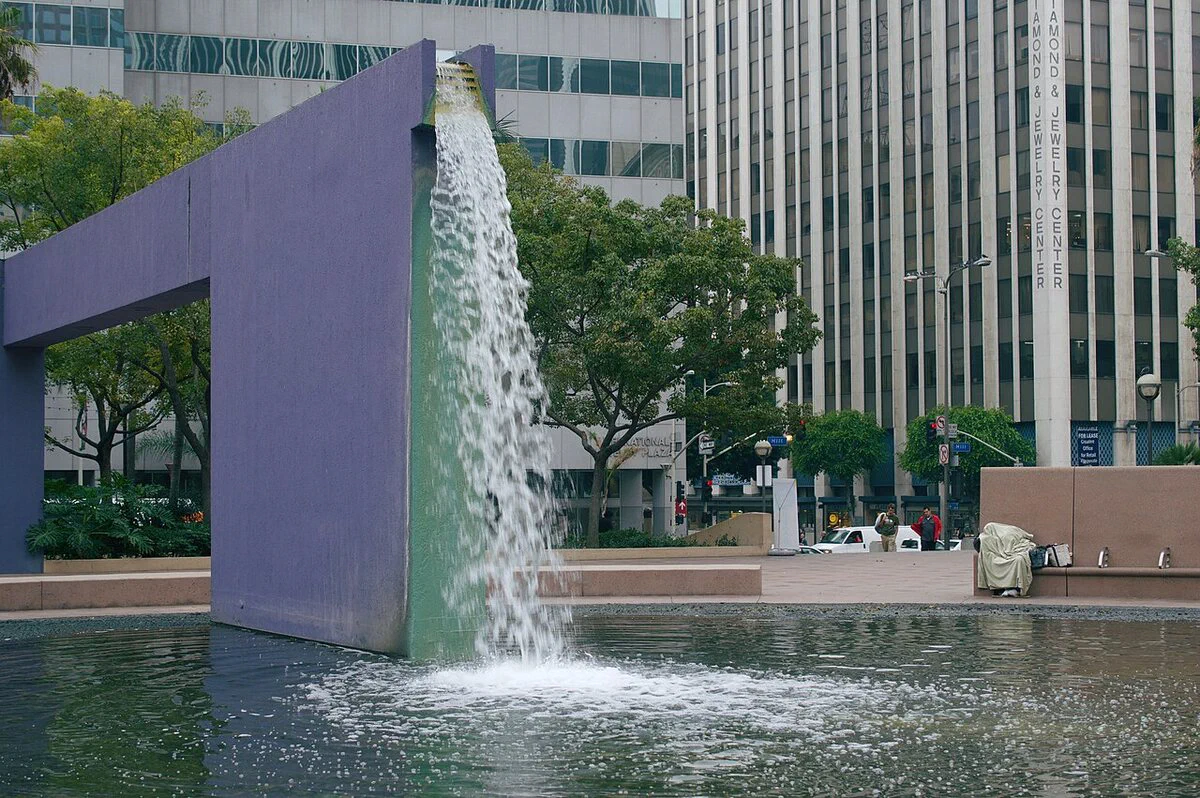 A purple concrete fountain wall and water basin at Pershing Square with downtown Los Angeles office towers behind